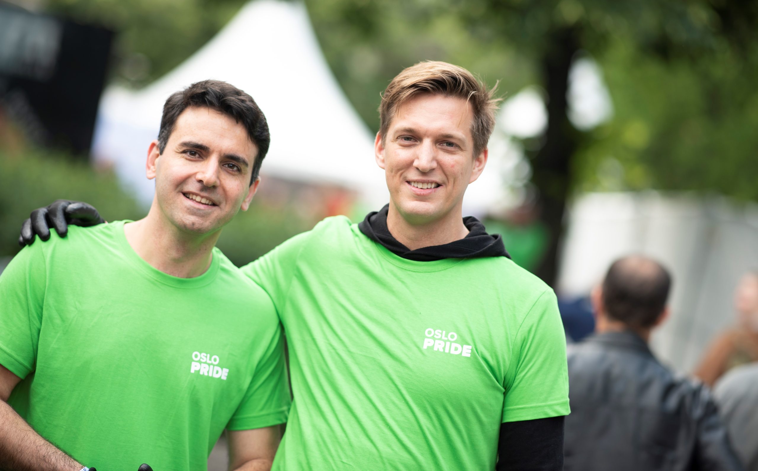 To hvite menn smiler til kamera. De har på seg grønne t-skjorter der det står Oslo Pride med hvit skrift. Den ene holder en arm rundt skulderen til den andre. Begge har sorte arbeidshansker på. De står foran noen hvite telt og sommergrønne trær, som vi skjønner er festivalområdet.