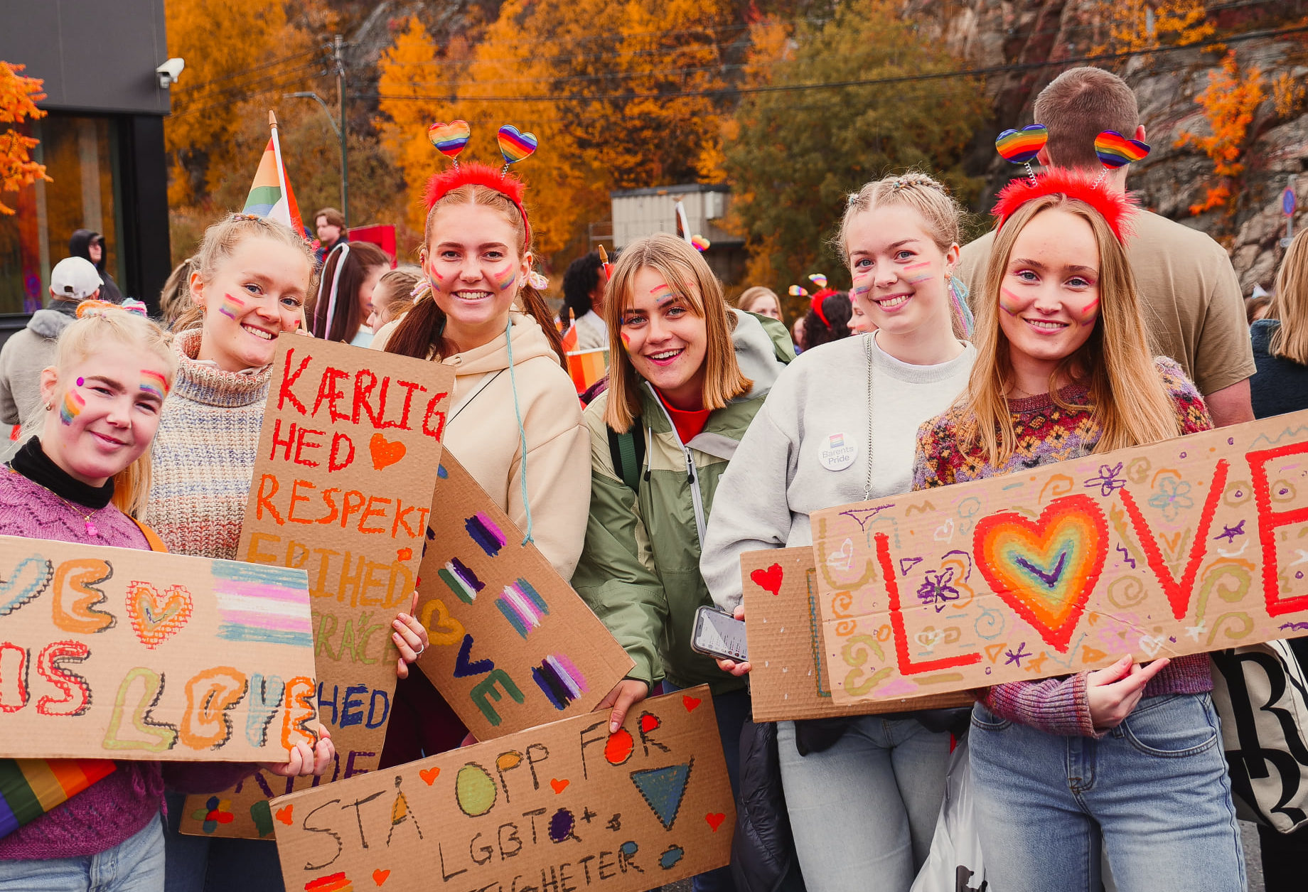 Seks ungekvinner står på rekke og smiler til kamera. De holder fargerike plakater med budskap om kjærlighet. og regnbuefarger. De har pyntet seg med prideeffekter. Bak dem er flere mennesker og trær med gult løv.