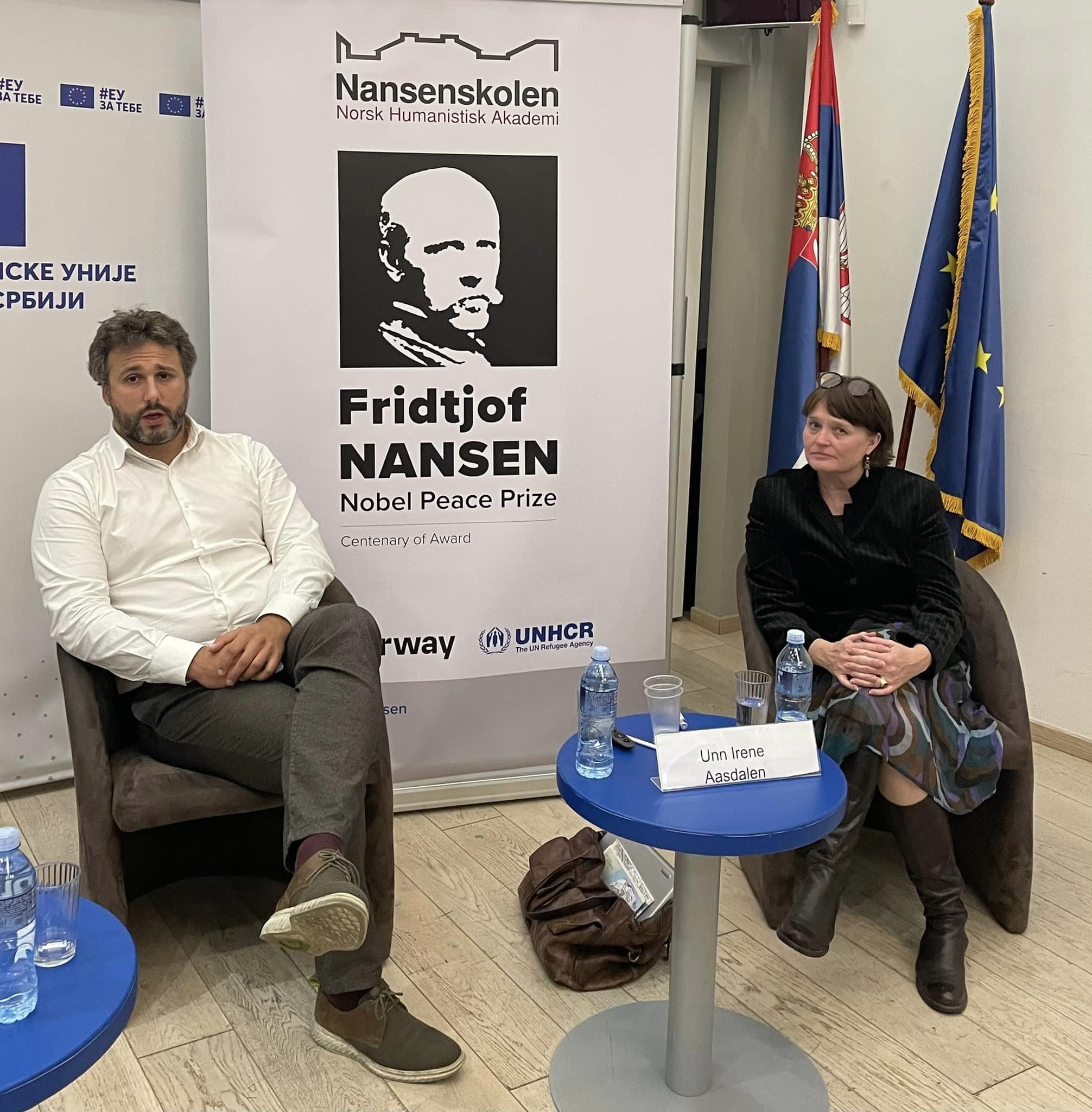 En hvit kvinne og en hvit mann sitter i hver sin stol foran en Rollup med bilde av Fridtjof Nansen. Bak kvinnen står to flagg, det ene er EU-flagget.  