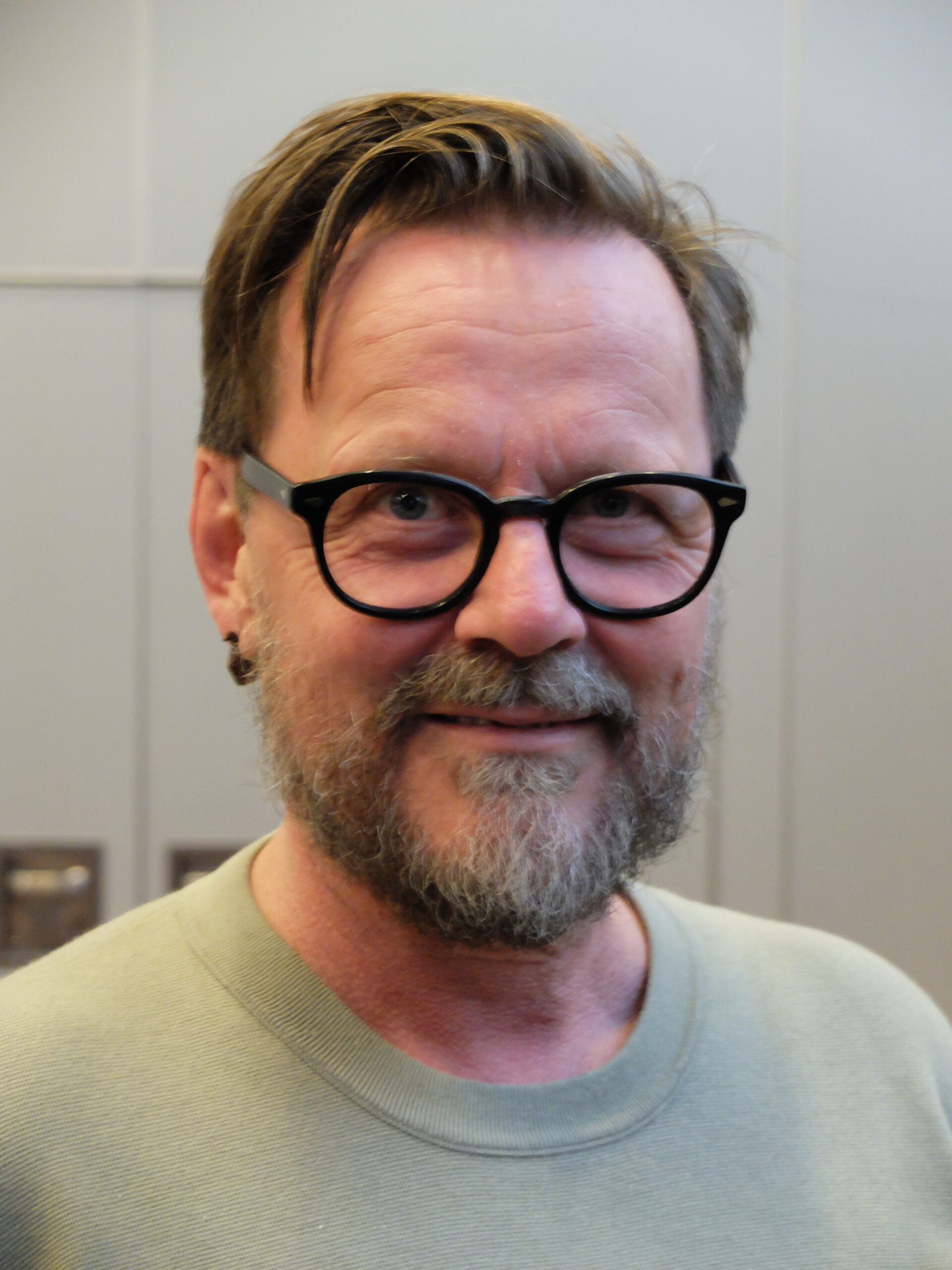 En hvit, voksen mann med skjegg, kort mørkt hår og briller, ser i kamera og smiler med øynene.