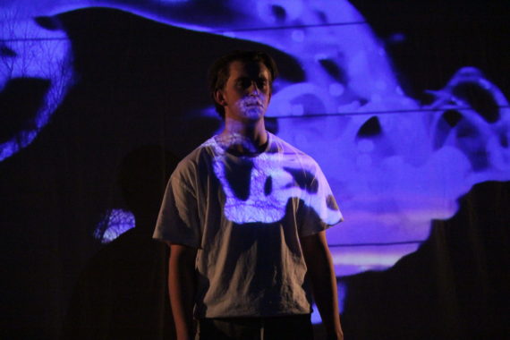 Person står i mørket med blått lys reflketert på deler av overkroppen og bakgrunnen. Vio forstår at hen står på en slags scene.