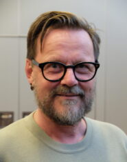 En hvit, voksen mann med skjegg, kort mørkt hår og briller, ser i kamera og smiler med øynene.